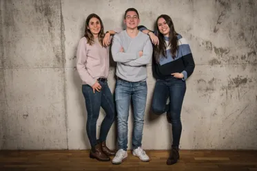 A relaxed group photo of three individuals standing against a textured concrete wall. They are casually dressed, two in sweaters and one in a light shirt, with arms crossed or rested casually. The setting features a warm, rustic wooden floor.